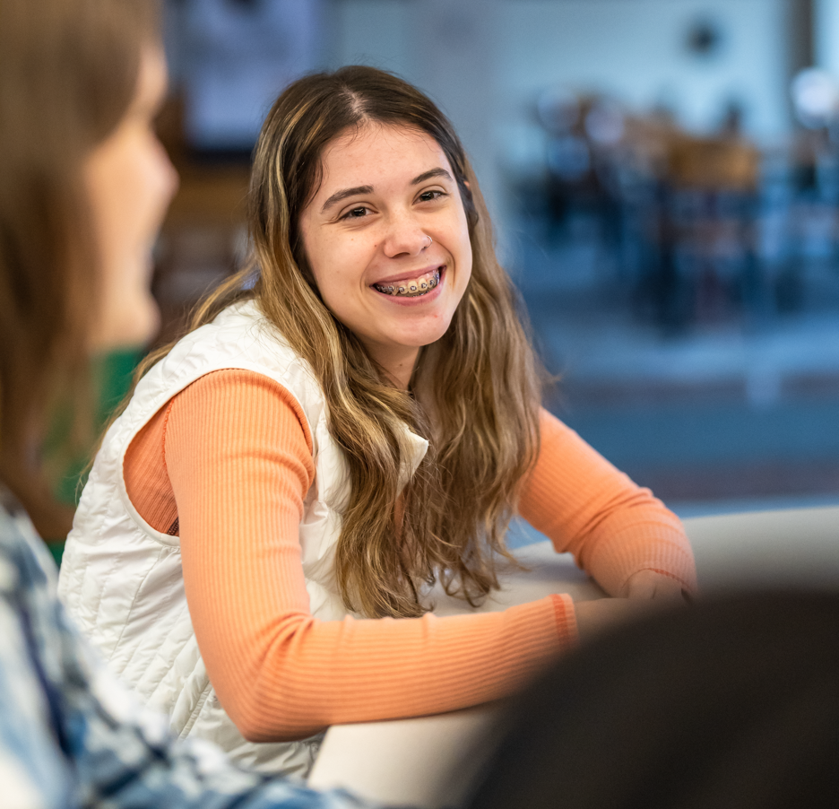 A student wearing a peach colored shirt and white vest is smiling and talking to other students sitting at a table in a study area.