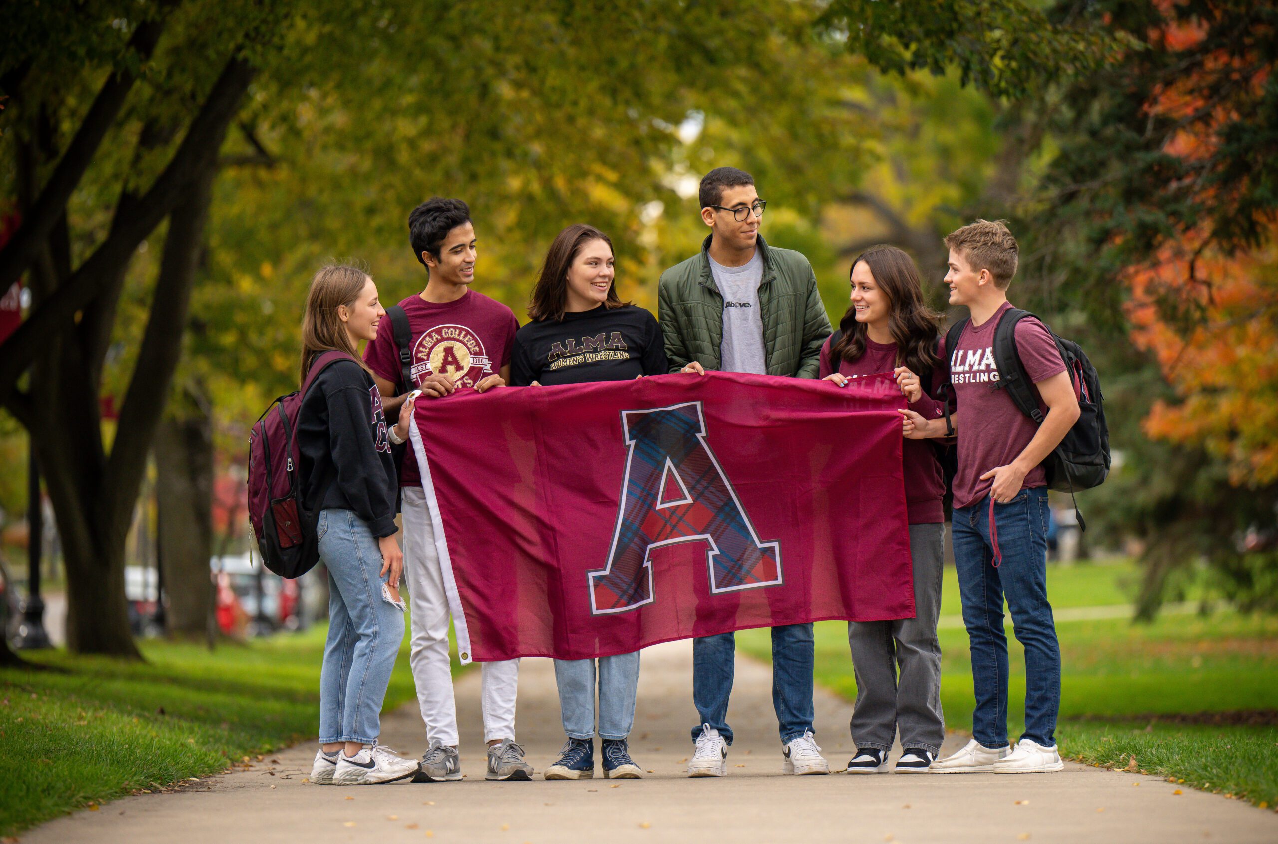 Six students standing on a sidewalk wearing Alma gear and holding an Alma College flag while exchanging glances and smiling at each other.