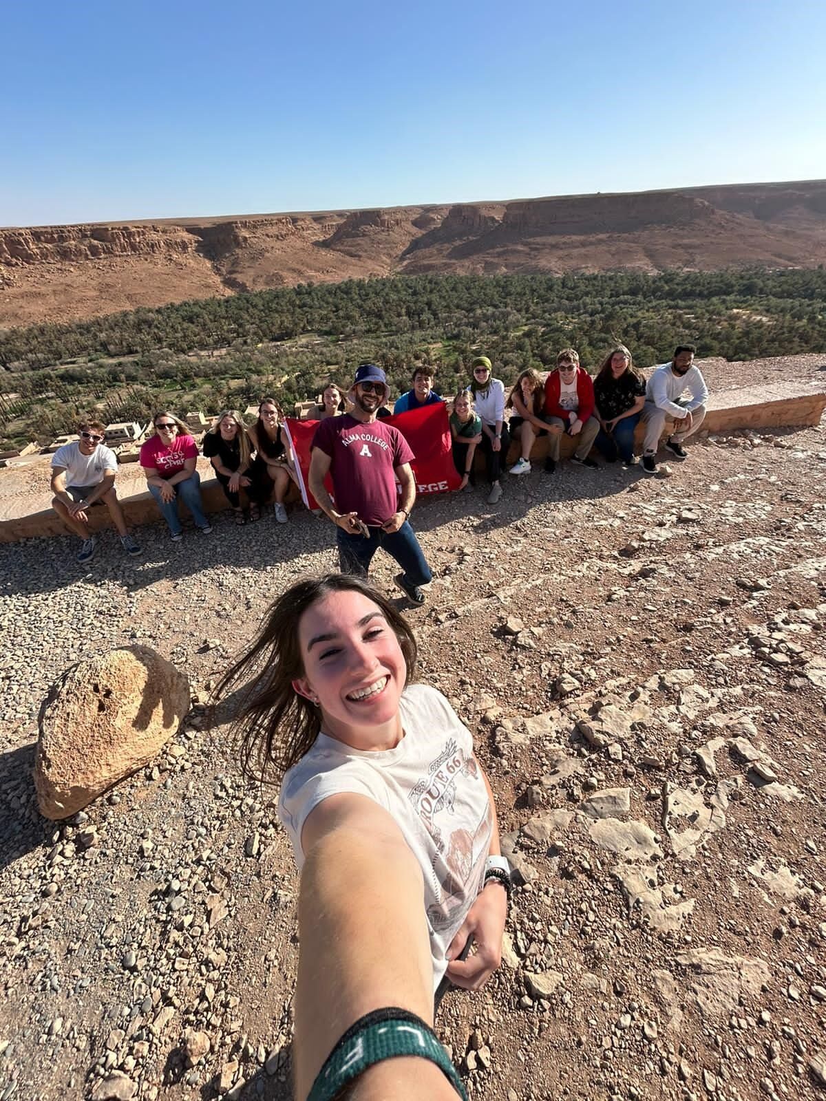 Student taking a selfie during a spring term course with the rest of the class sitting behind her and posing for the camera with the Alma College flag with canyon-scapes in the background.