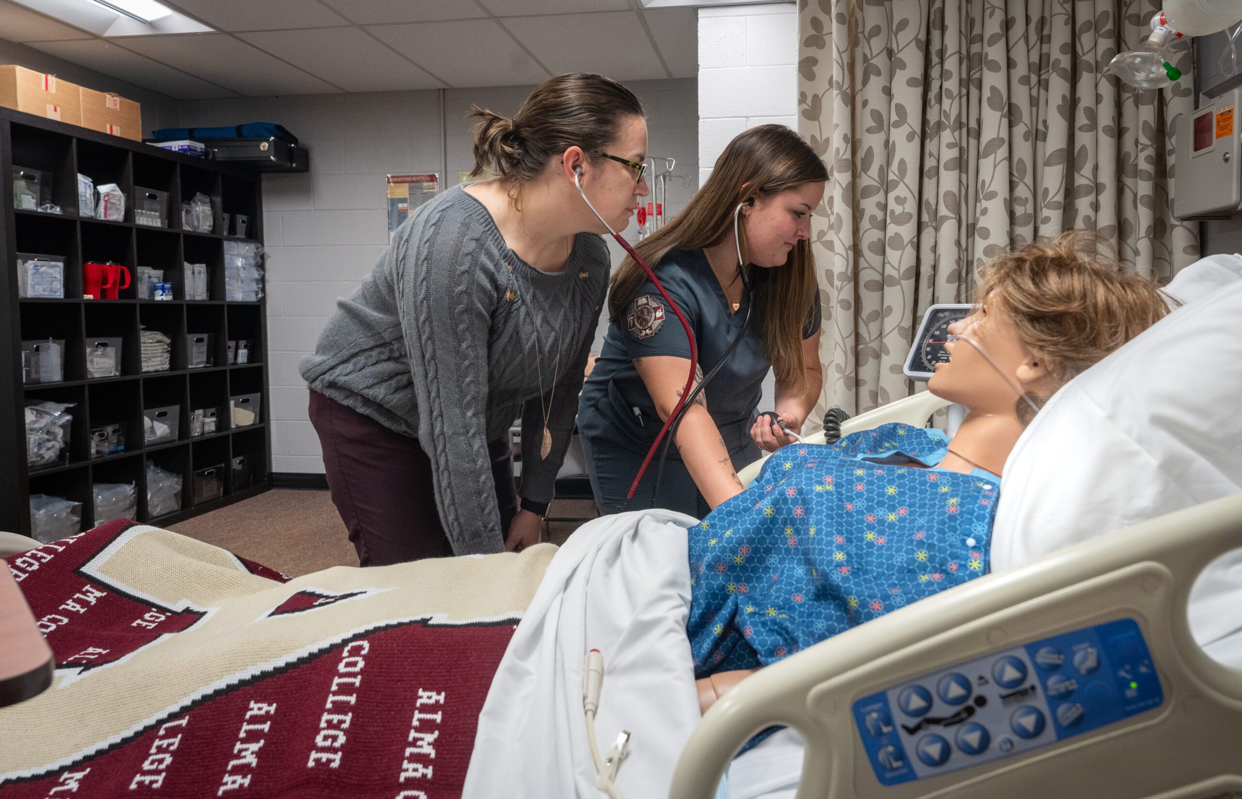 a nursing student and instructor taking blood pressure and listen in their stethoscope as the student trains on a manikin in the Nursing Skills Sim Lab on the campus of Alma College.