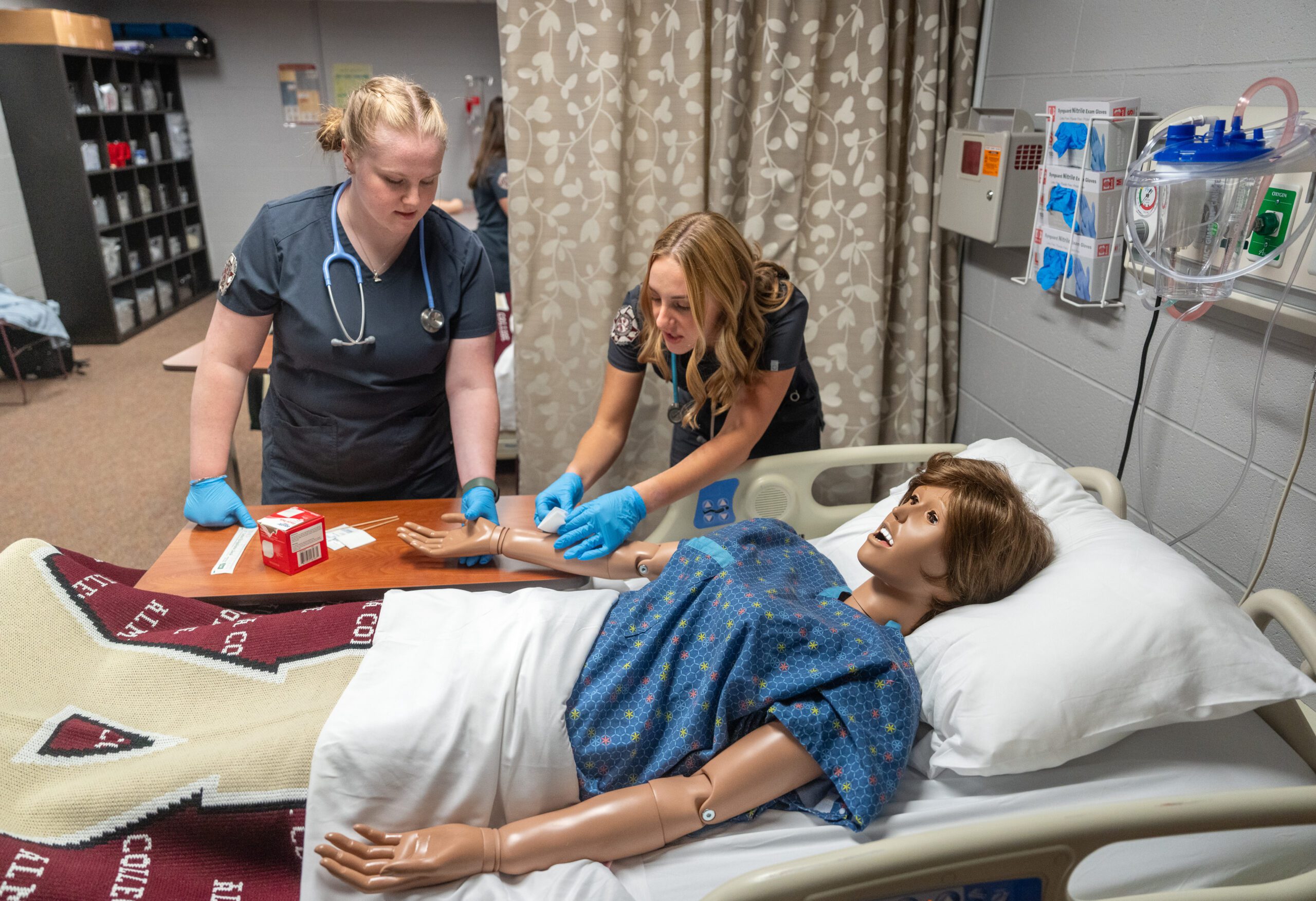 Two nursing students tending to the forearm of a training manikin in the Nursing Skills Sim Lab at Alma College