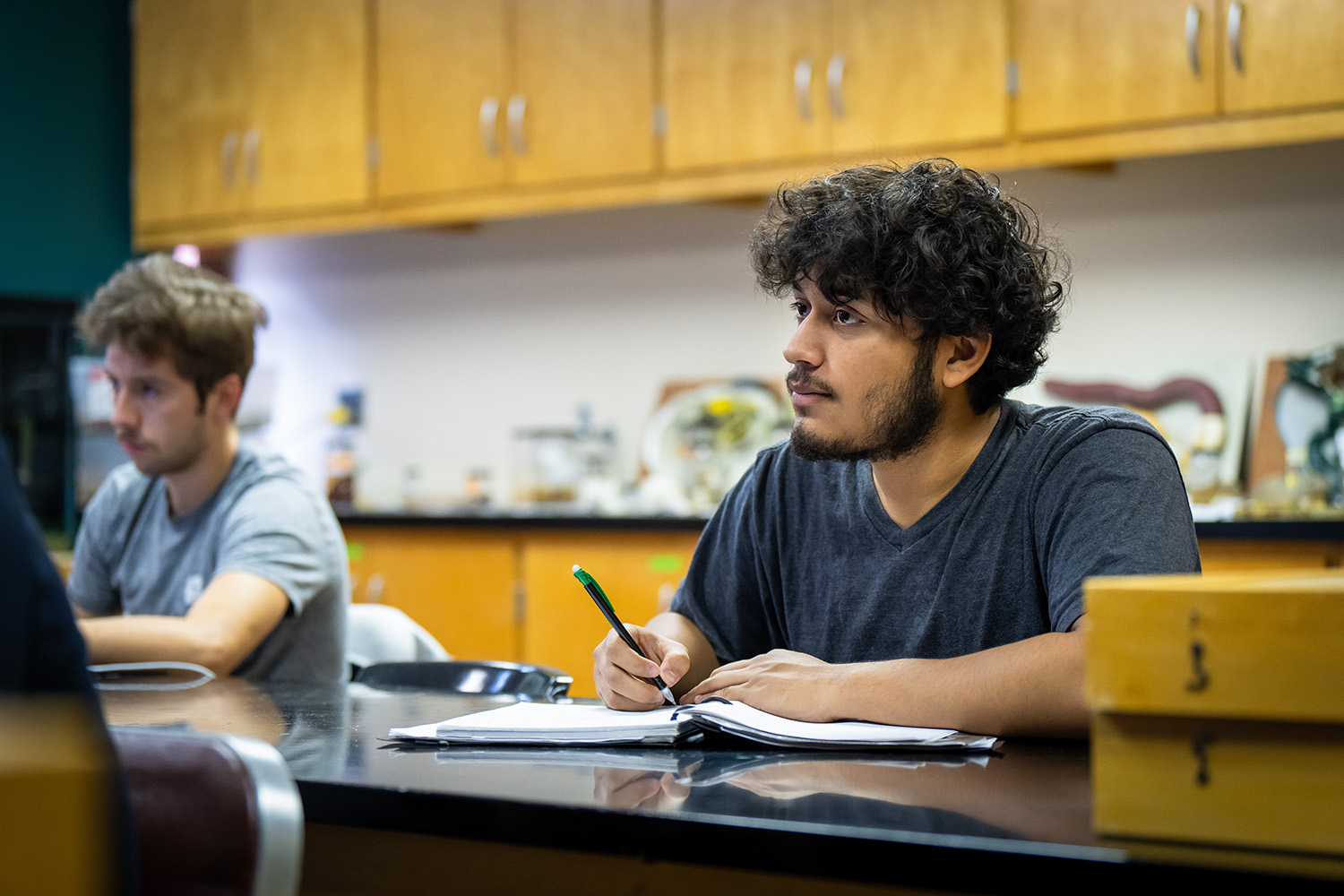 Student sitting at a table in a lab-style classroom with their notebook open and writing notes.