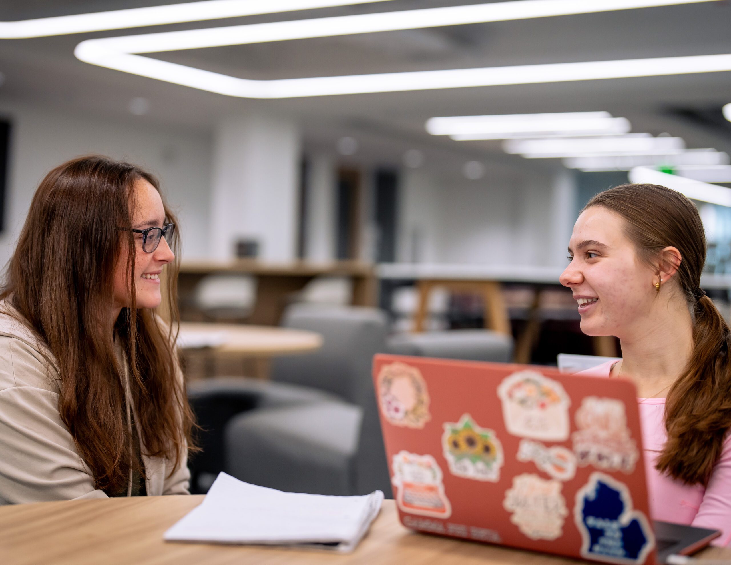 Two students outside of the Center for Campus and Community Engagement smiling while having a conversation at a table that has an open laptop and papers on top.