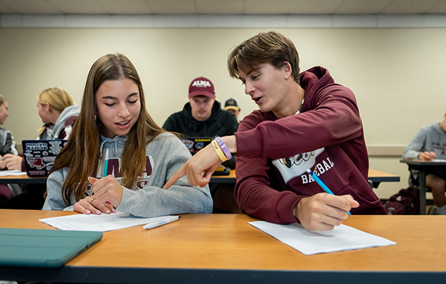 Two students sitting next to each other in class talking and working on a project together.