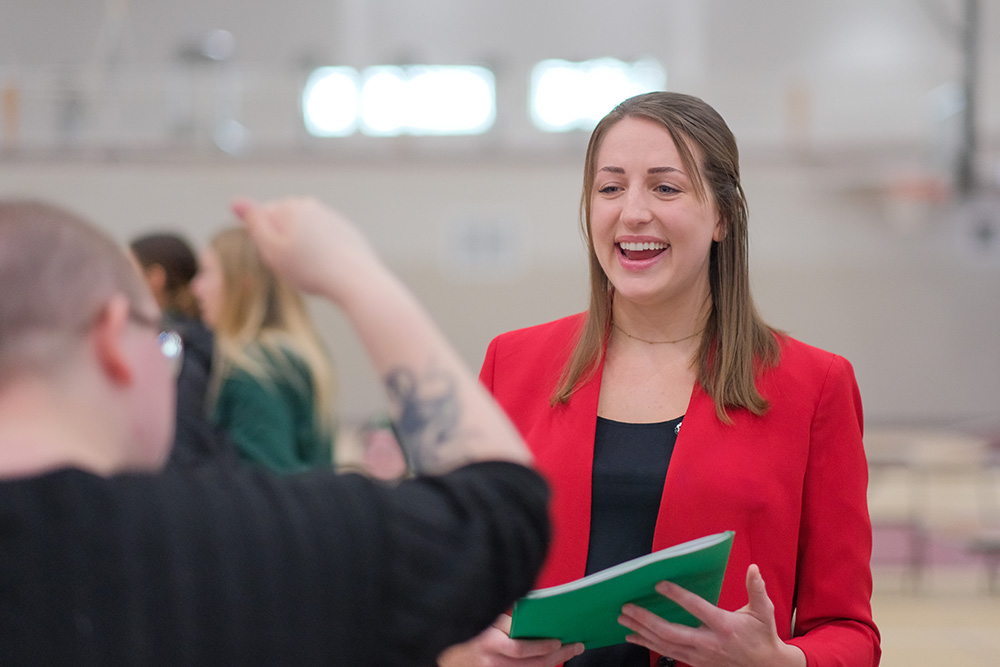 A female student wearing professional clothing and holding a folder talking to an employer at Alma College