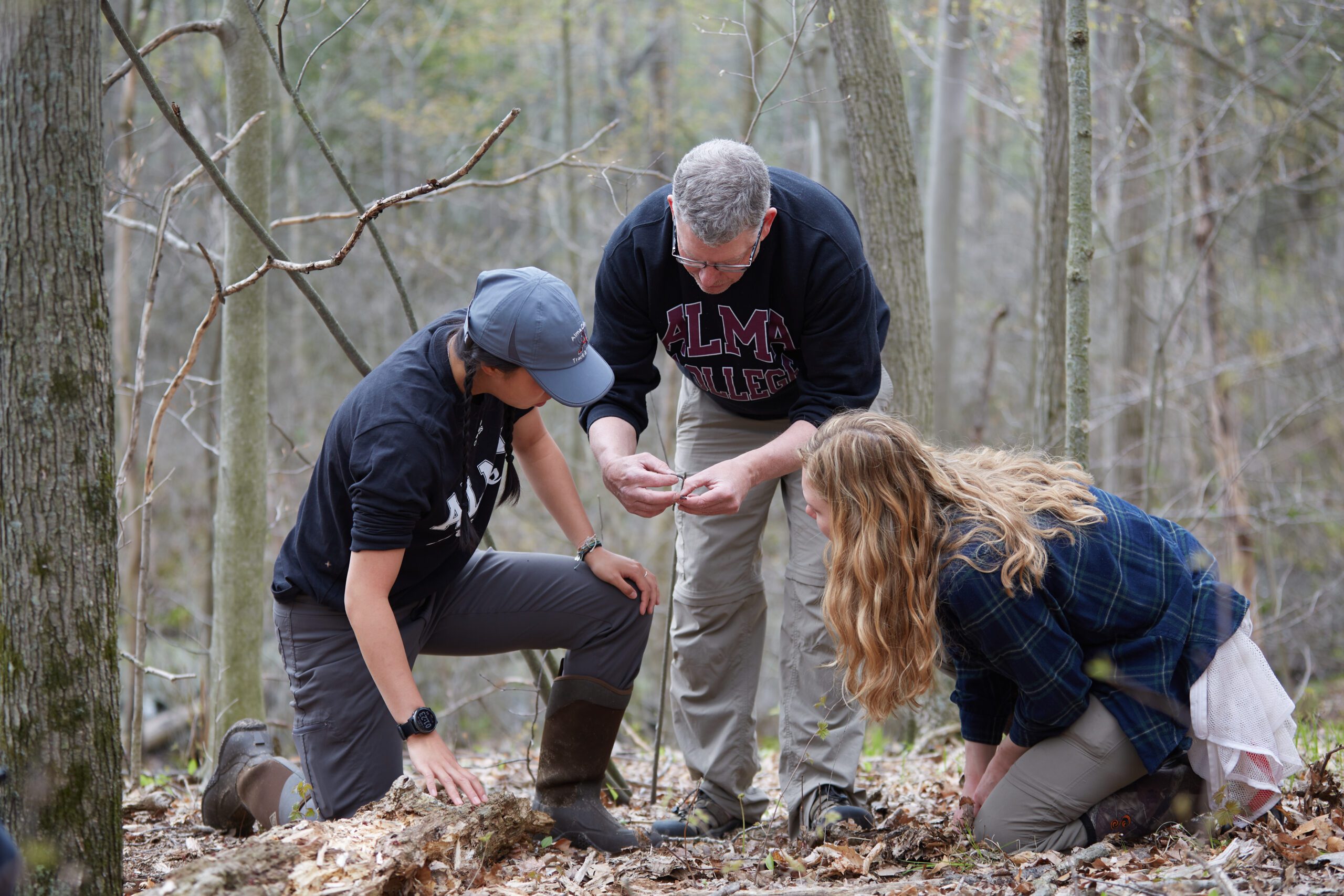 Photo of two students and a professor working outside and the professor is holding something in their hands that both students are looking at.