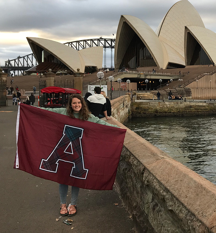 Student standing and holding the Alma College Flag in front of the Sydney Opera House in Sydney Australia.