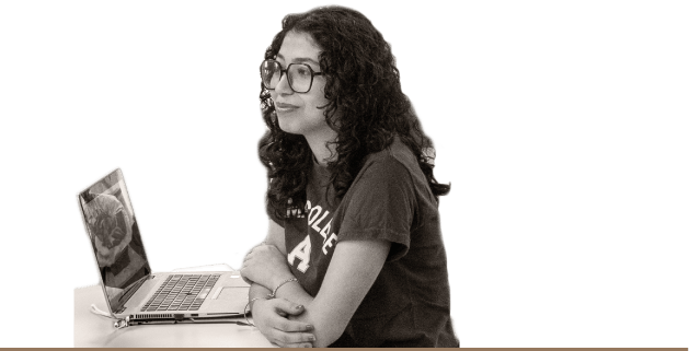 A woman wearing glasses sits at a desk, focused on her laptop, surrounded by a cozy workspace.